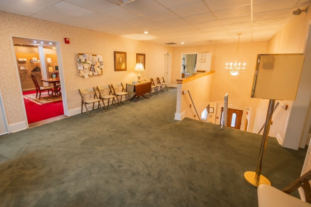 A funeral home interior. Green carpeted hallway with chairs, stairs, and a chandelier, with a room visible to the left.