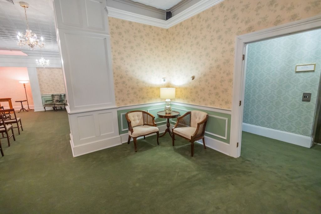 A well-lit room with two chairs by a table, set on green carpet. A doorway is visible to the right.