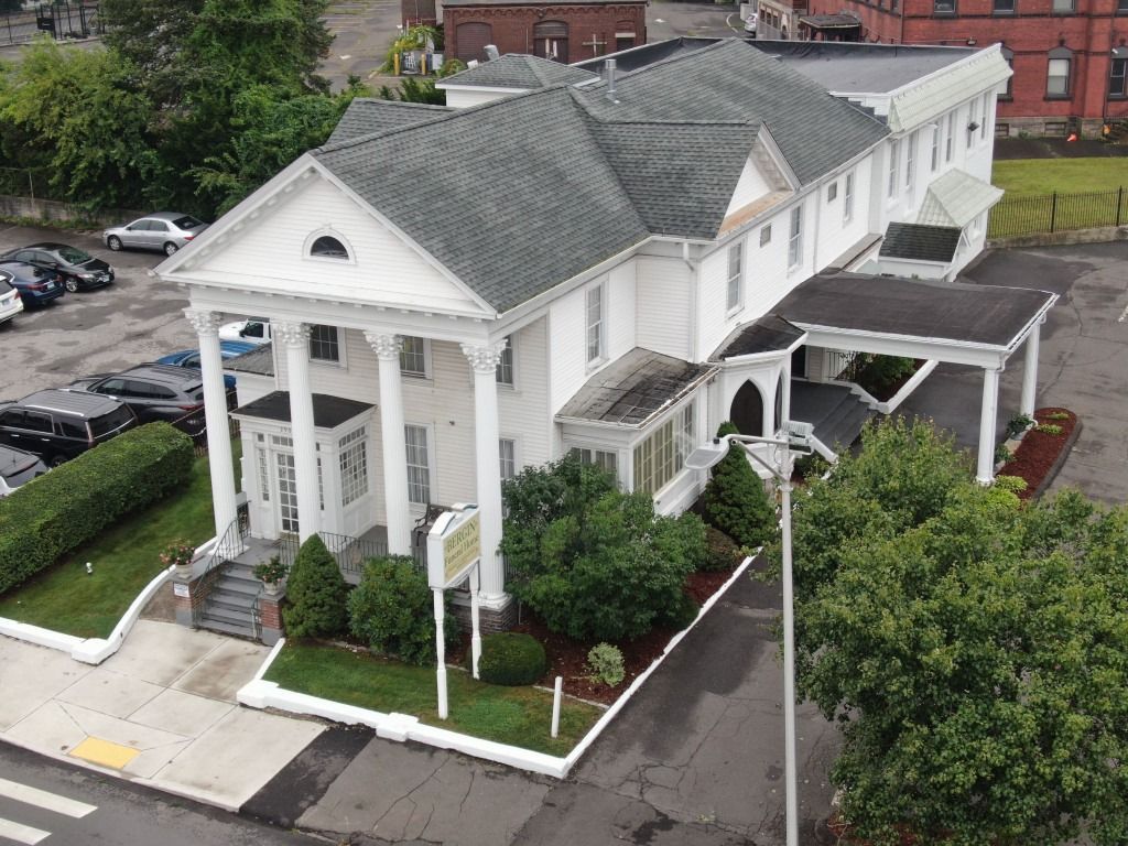 White funeral home building with pillars, driveway, cars, and landscaping.