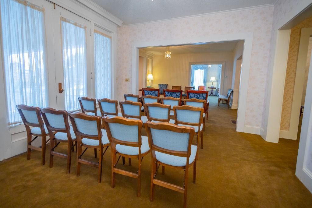 Empty room with rows of chairs facing a door, light blue chair cushions, windows with curtains, and carpet.