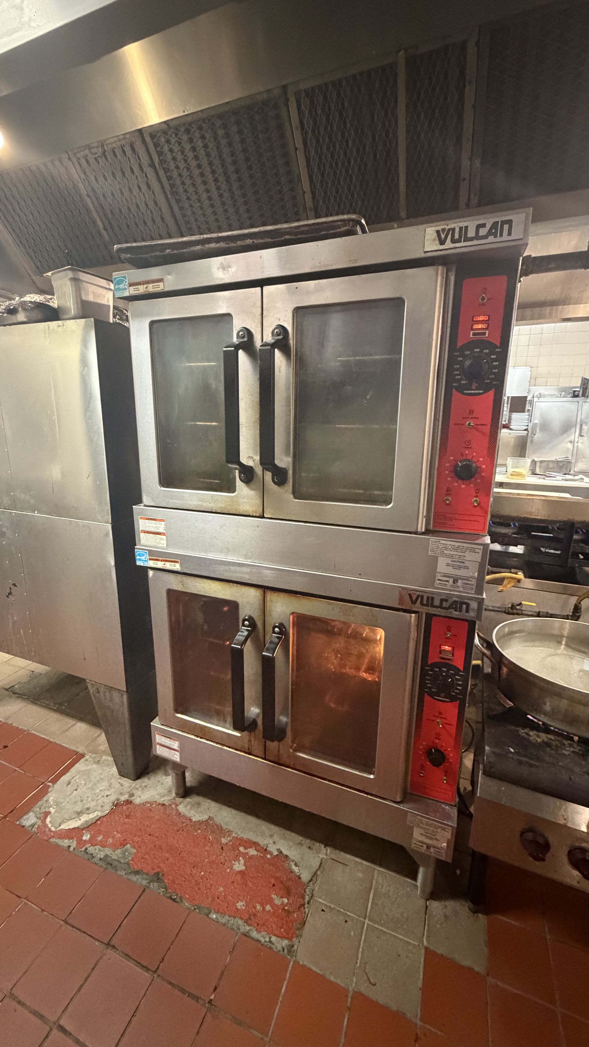 Stainless steel commercial oven with black handles in a kitchen, surrounded by red tile floor.