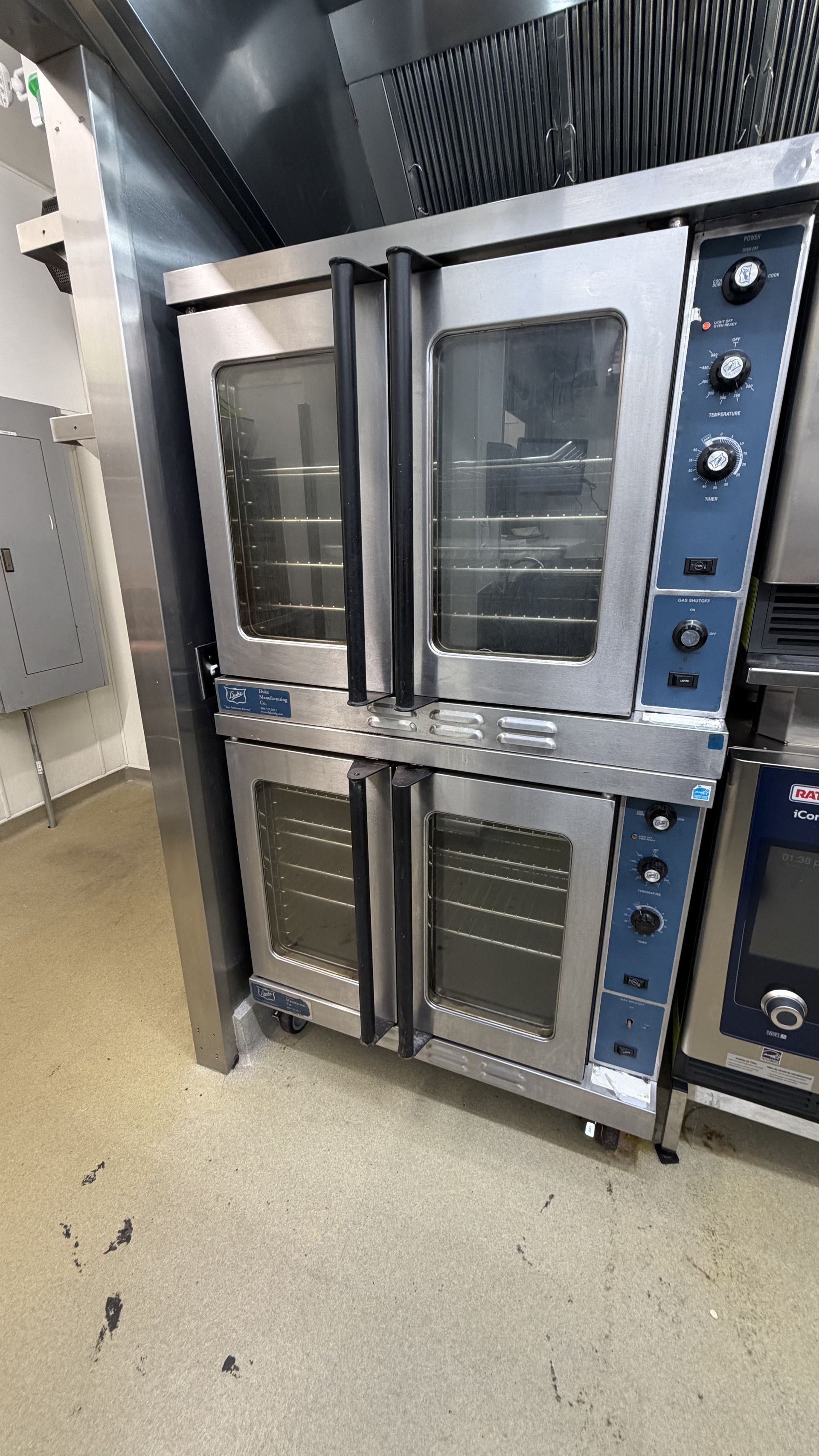Stacked stainless steel commercial ovens in a kitchen, one door open, on a tiled floor.