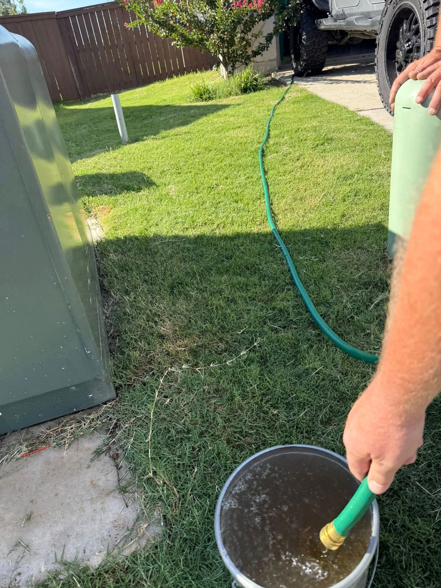 Person watering a lawn with a green hose and bucket near a house and parked car