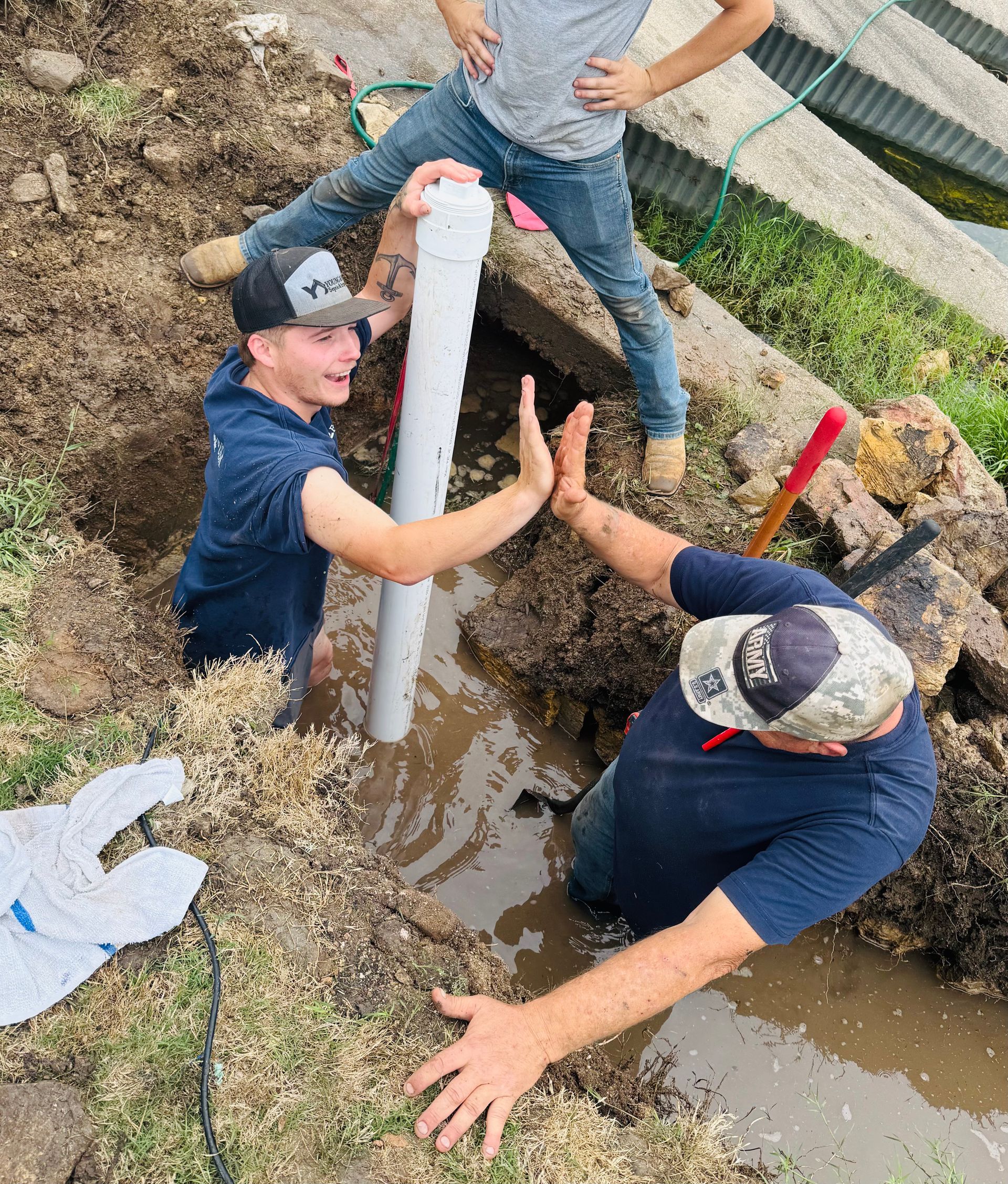 Workers installing a white pipe in a muddy trench beside a fence