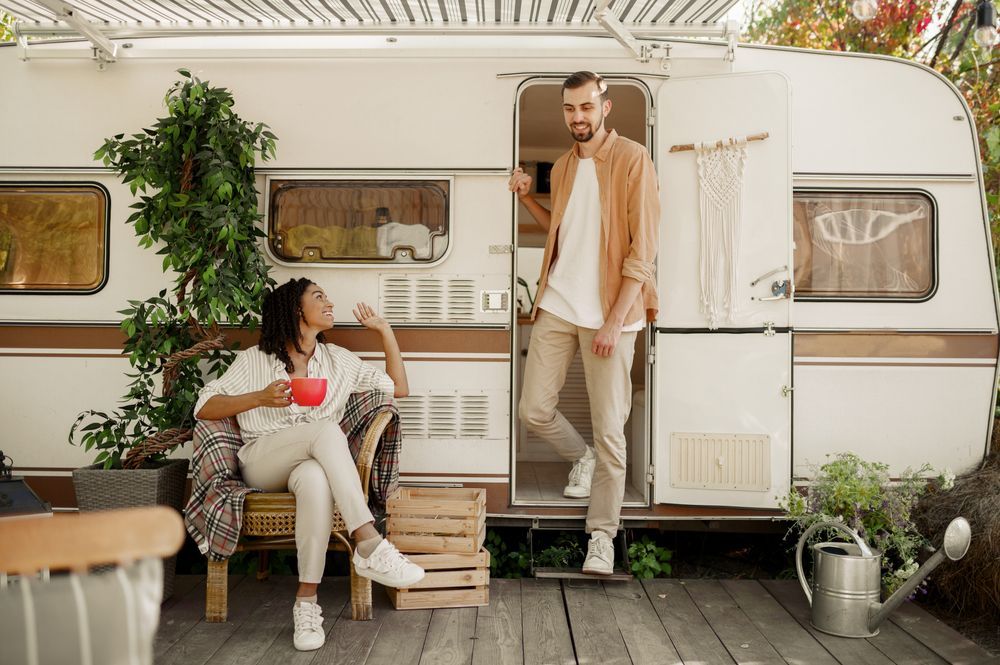 Couple relaxing by a vintage camper on a deck; woman seated, holding a mug; man leaning in the doorway.