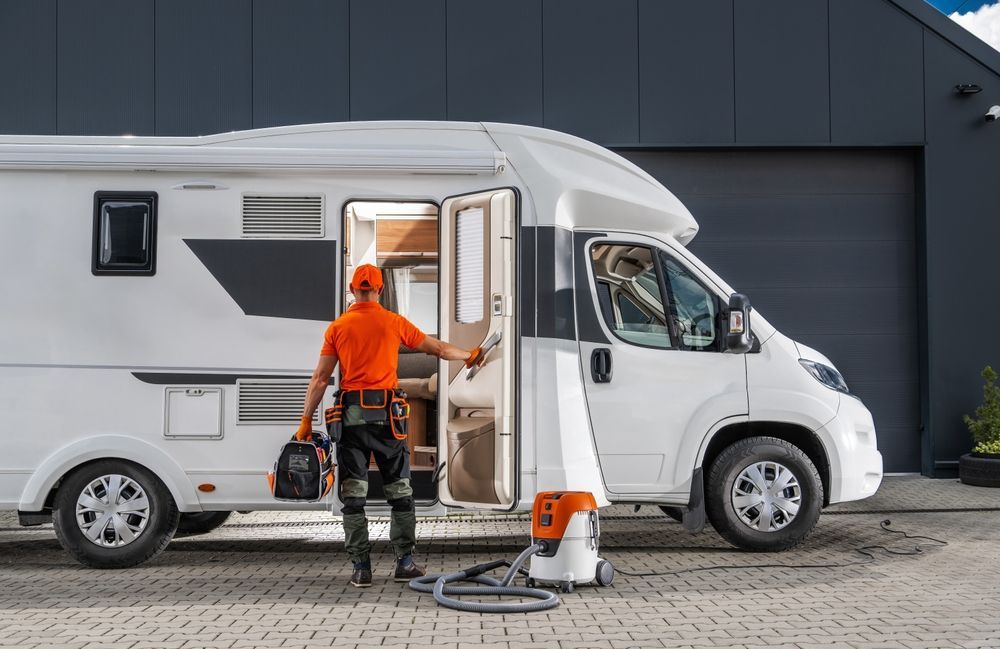 Man with tools enters a white RV. A vacuum cleaner sits by the open door. Garage in the background.