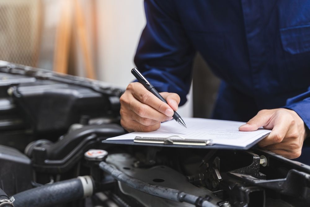 Mechanic in blue jumpsuit writing on a clipboard while inspecting a car engine.