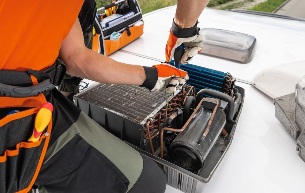 HVAC technician repairing an air conditioning unit; wearing gloves, on a rooftop. Orange shirt, tool bag visible.