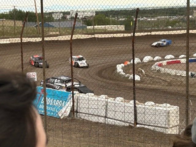 Race cars racing on a dirt track, viewed through a fence. Cars are near the curve, with spectators in the foreground.