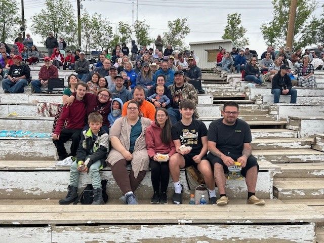 Group of people, mostly adults and children, seated in bleachers at an outdoor event, some smiling.