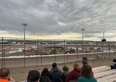 Spectators watch a dirt track race under a cloudy sky. Trucks and vehicles are on the track.