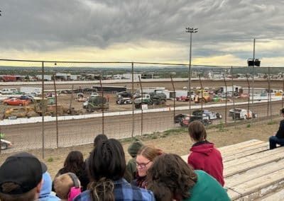 People watch a dirt track race from bleachers, cars on track, cloudy sky.