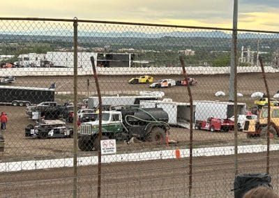 Race cars on a dirt track, viewed through a chain link fence. Trucks, trailers, and buildings in the background.