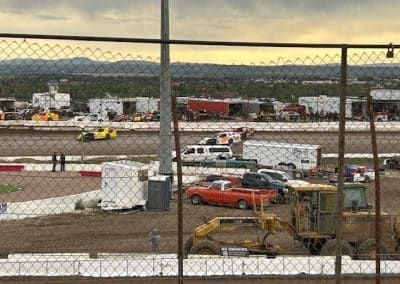 Race cars on a dirt track, viewed through a chain-link fence. Other vehicles and equipment are also in view.