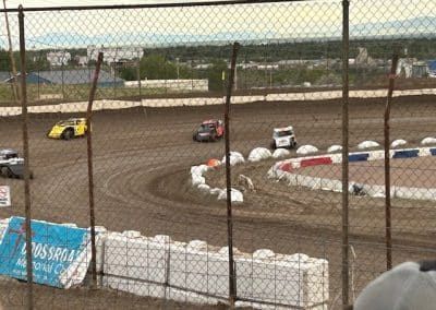 Race cars racing on a dirt track, near white safety barriers.