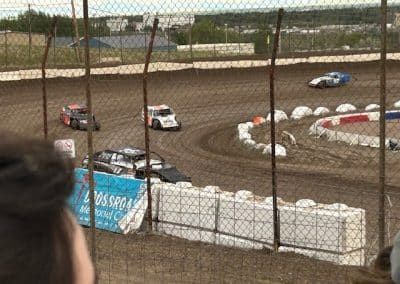 Dirt track race with cars on a curved track, viewed through a fence.