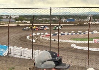 Race cars on a dirt track, viewed through a fence. Mountains visible in the background. Spectator in the foreground.
