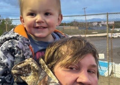A child smiles on a person's shoulders outdoors. Child wears camo jacket; person wears camo jacket.