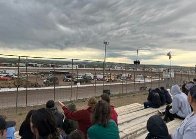 People watch a dirt track race under a cloudy sky.