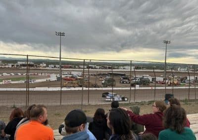 People watch a dirt track race under a cloudy sky. Cars race on a dusty track, behind a wire fence.