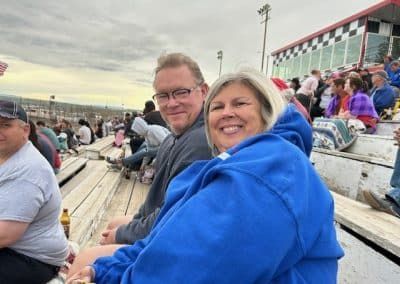 Couple smiling in bleachers at a racetrack; woman in blue hoodie, man with glasses, other spectators in the background.