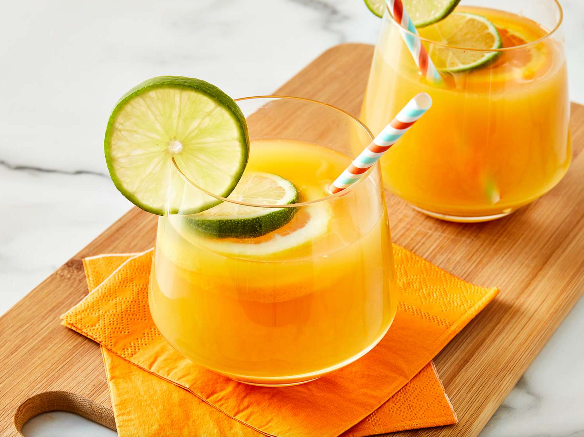 Two glasses of orange juice with straws and lime slices on a wooden cutting board.