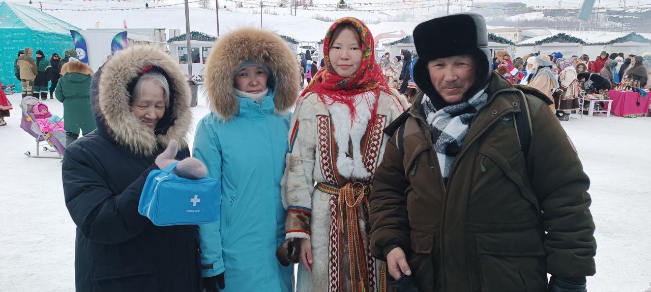 Four people in winter clothing posing outdoors at a snowy festival, with colorful tents in the background.