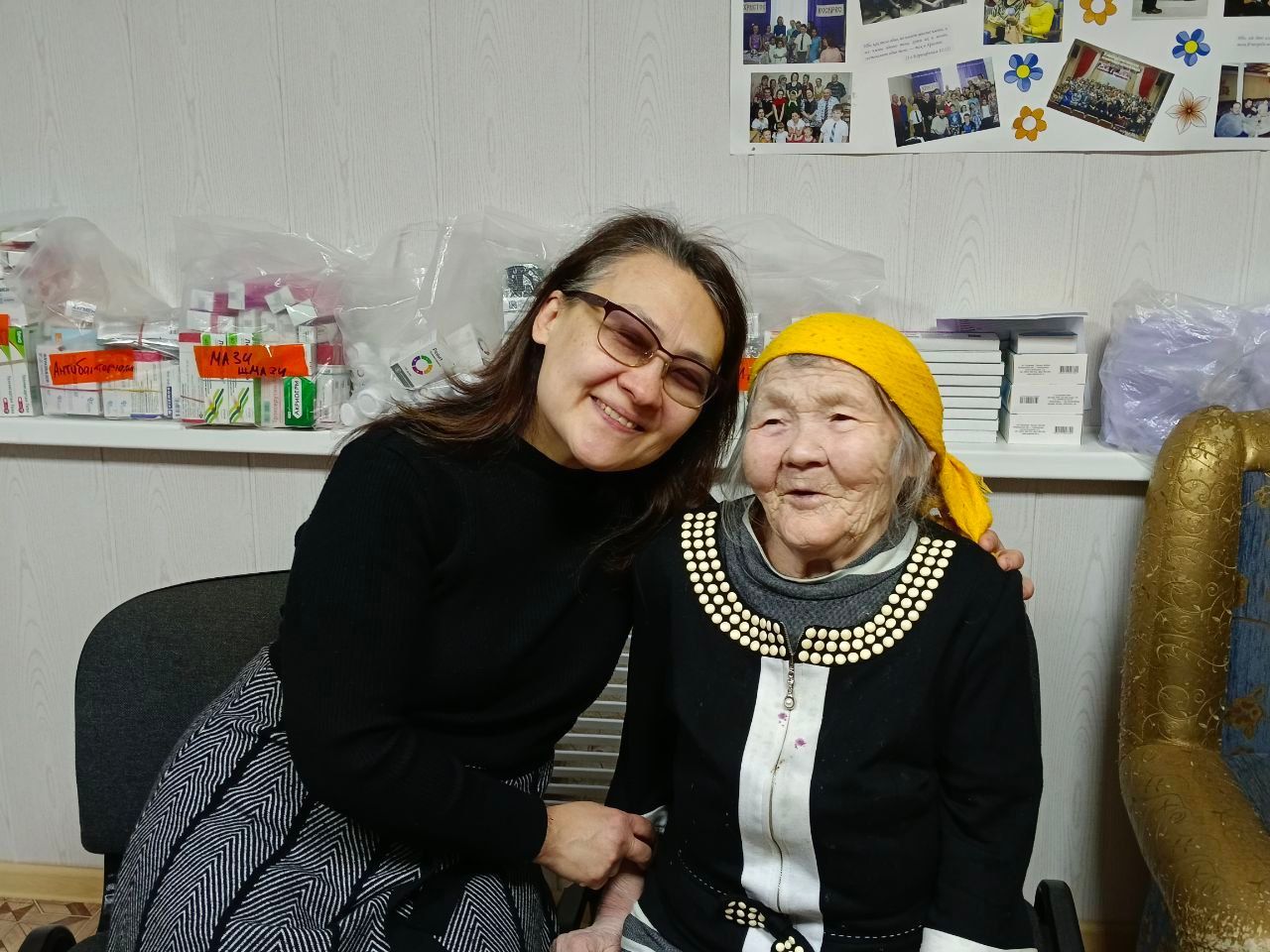 Two women sitting close together, smiling in a room with photos and shelves in the background