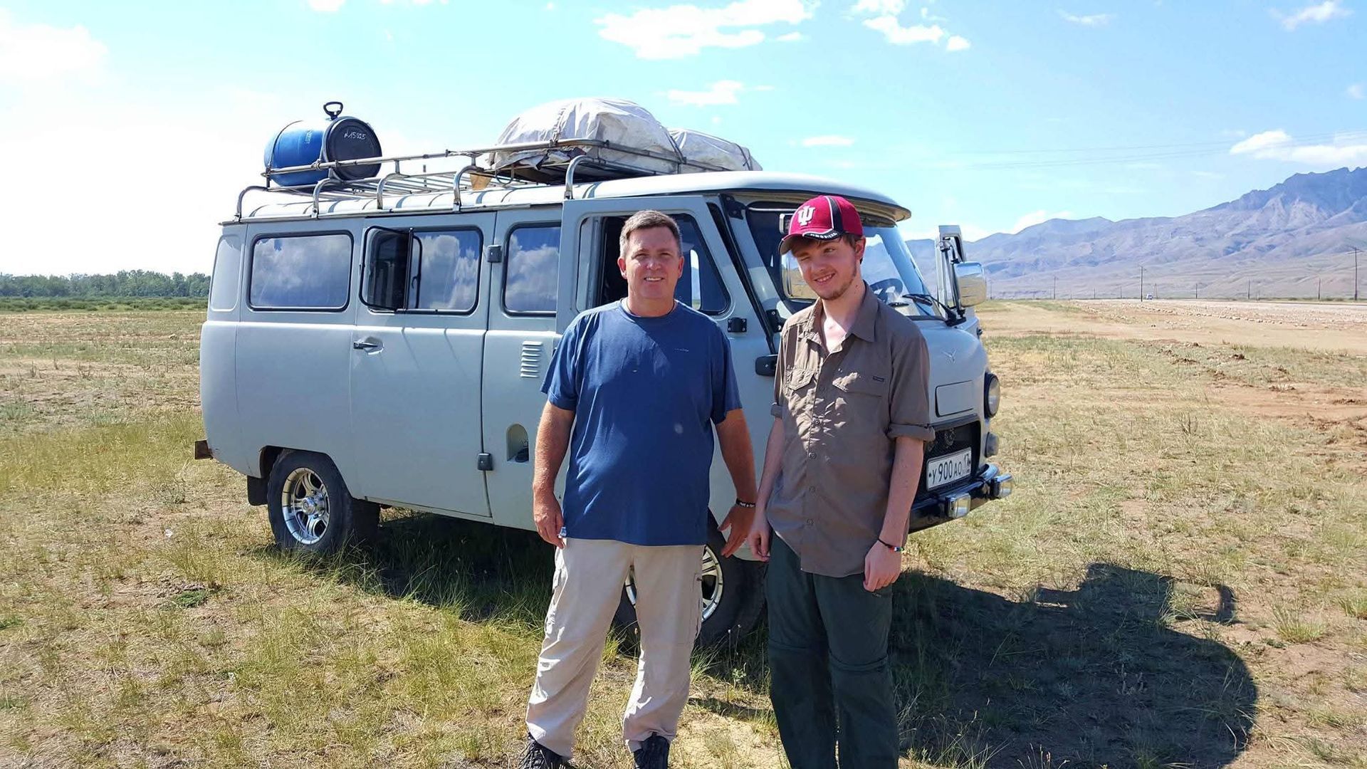 Two people standing beside a vintage van in a grassy field with mountains in the distance.