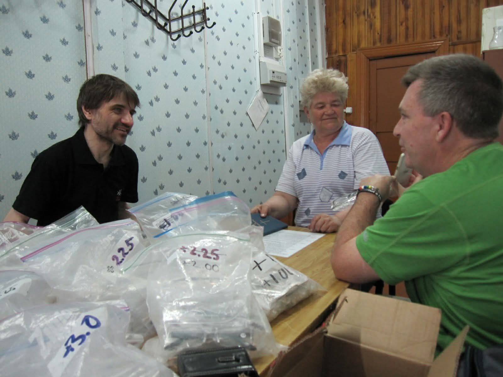 Three people sit at a table sorting plastic bags and boxes in a room with patterned wallpaper.