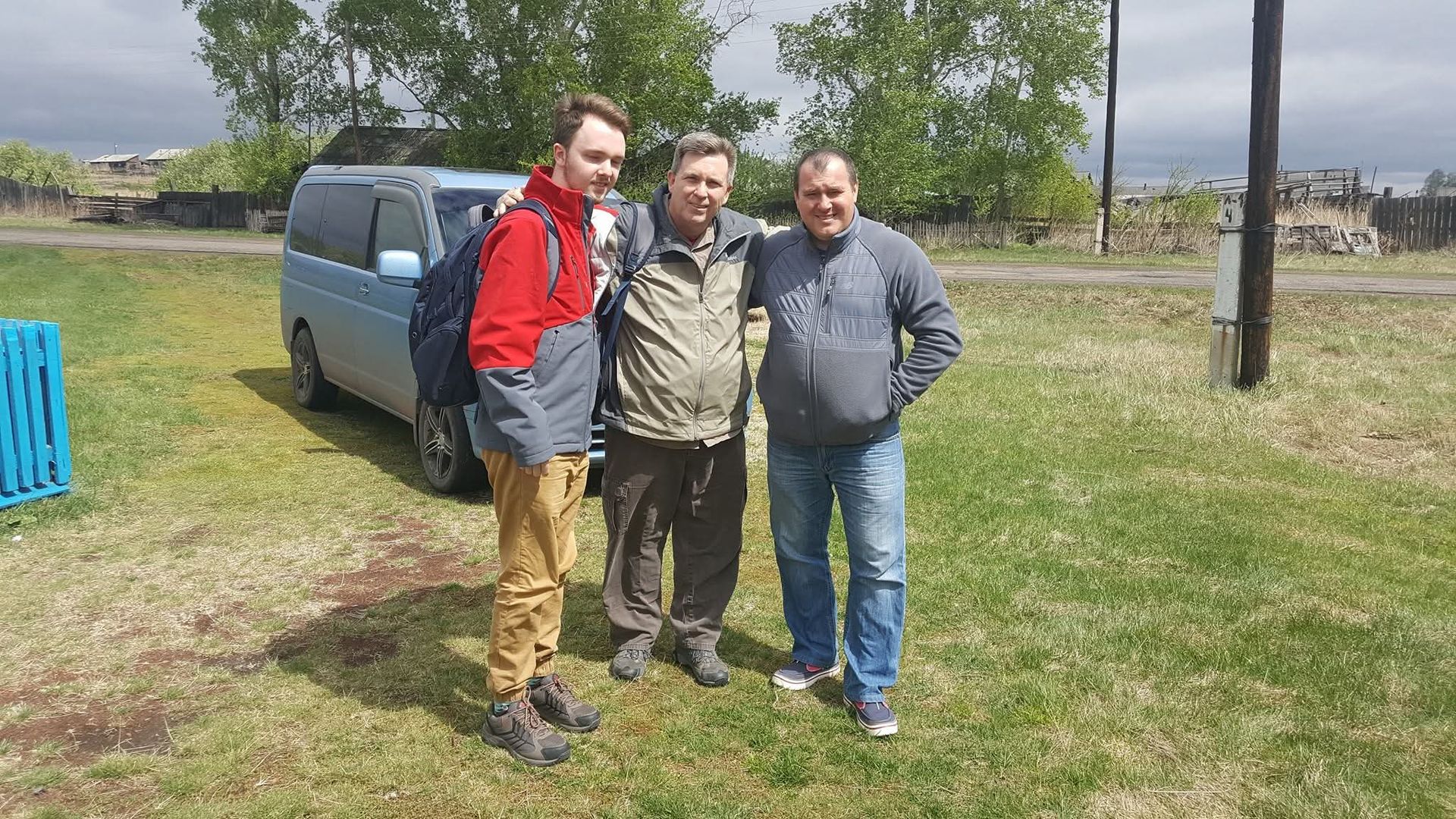 Three people standing together on a grassy field beside a blue van, with trees and open land behind them.