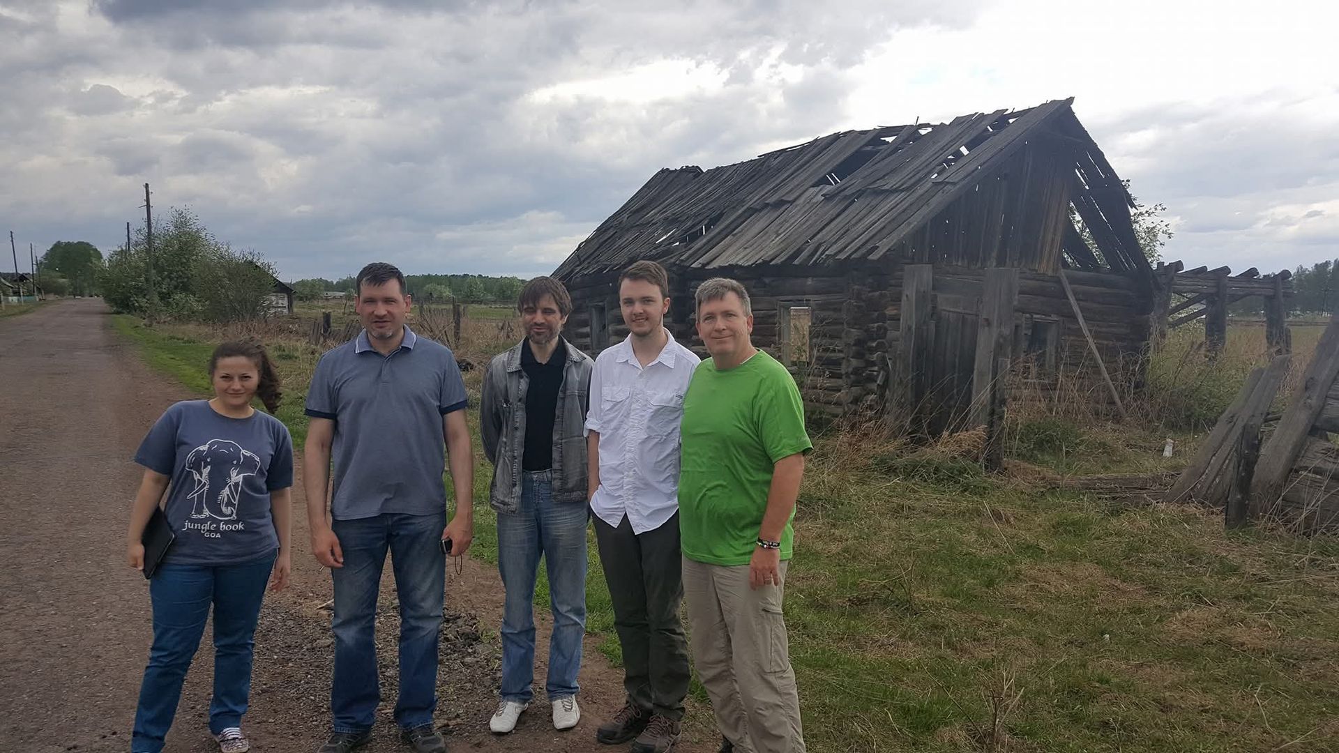 Five people stand in front of a burned-out house in a grassy field under a cloudy sky.