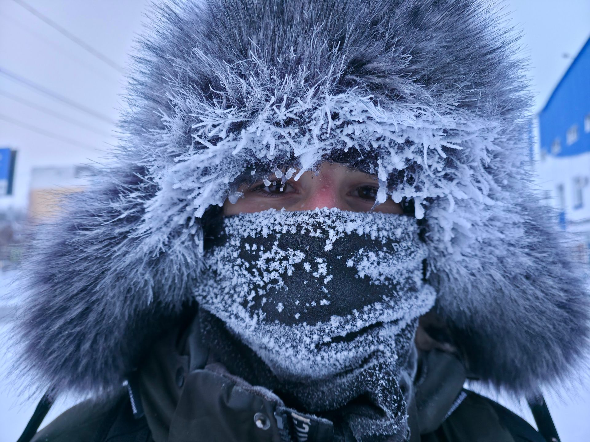 Person in a fur-lined hood and face covering, standing in snowy, icy weather.