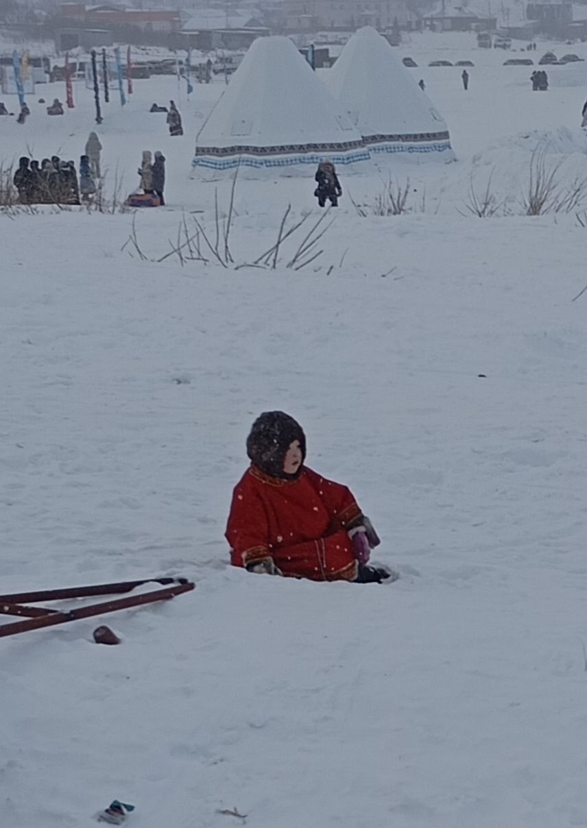 Child in a red jacket sitting in deep snow near sleds, with people and tents in a snowy background.