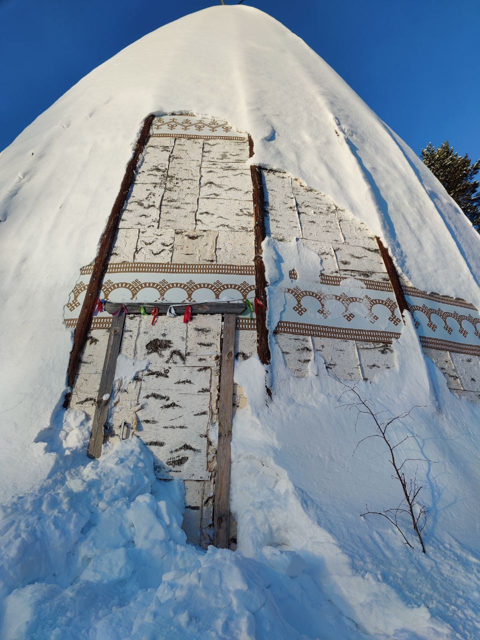 Snow-covered tall structure with a narrow, decorated doorway or ladder-like frame against a snowy hillside