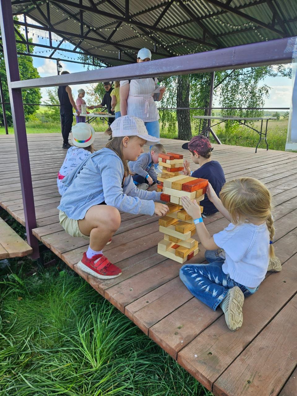 Children play giant Jenga on a wooden deck under a shaded outdoor shelter.