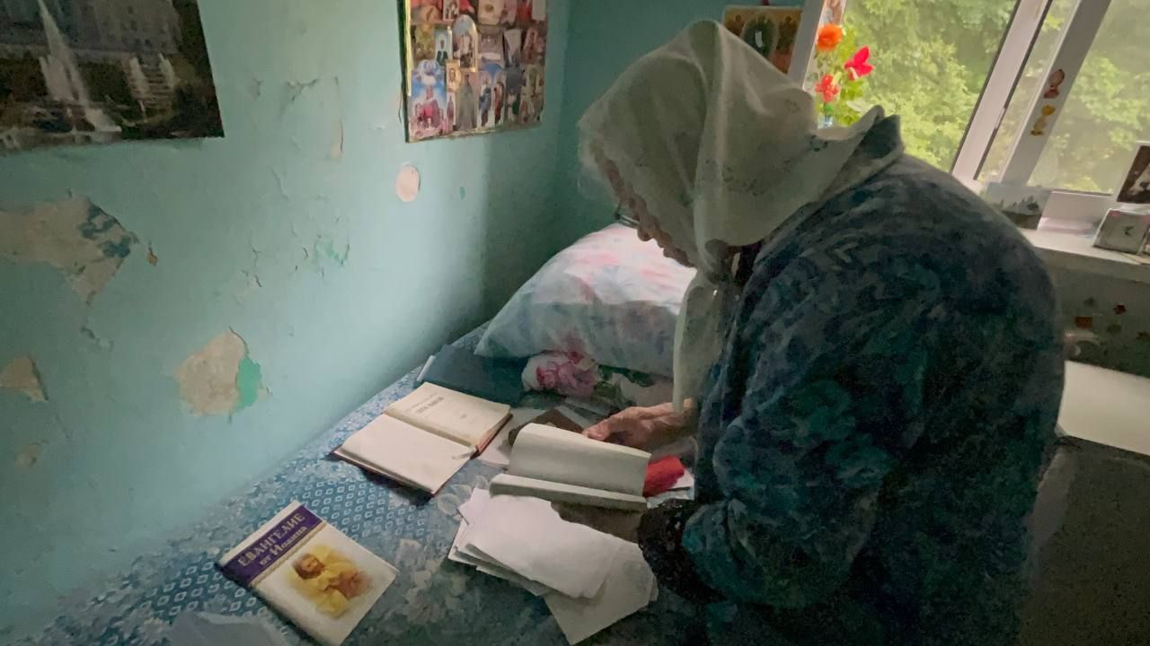 Person studying at a desk in a small bedroom with open books and papers by a window.