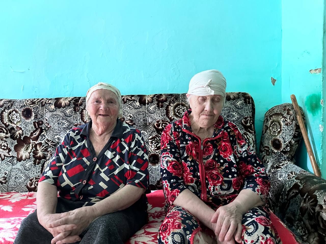 Two women sit on a patterned couch against a turquoise wall, wearing red and black floral clothing.