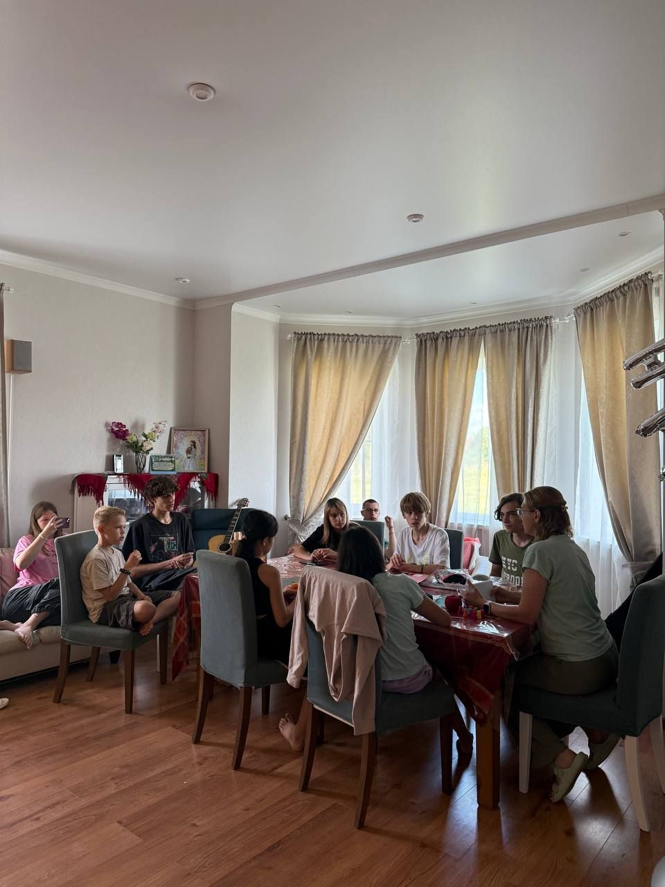 People seated around a dining table in a bright living room, sharing a meal near large windows.