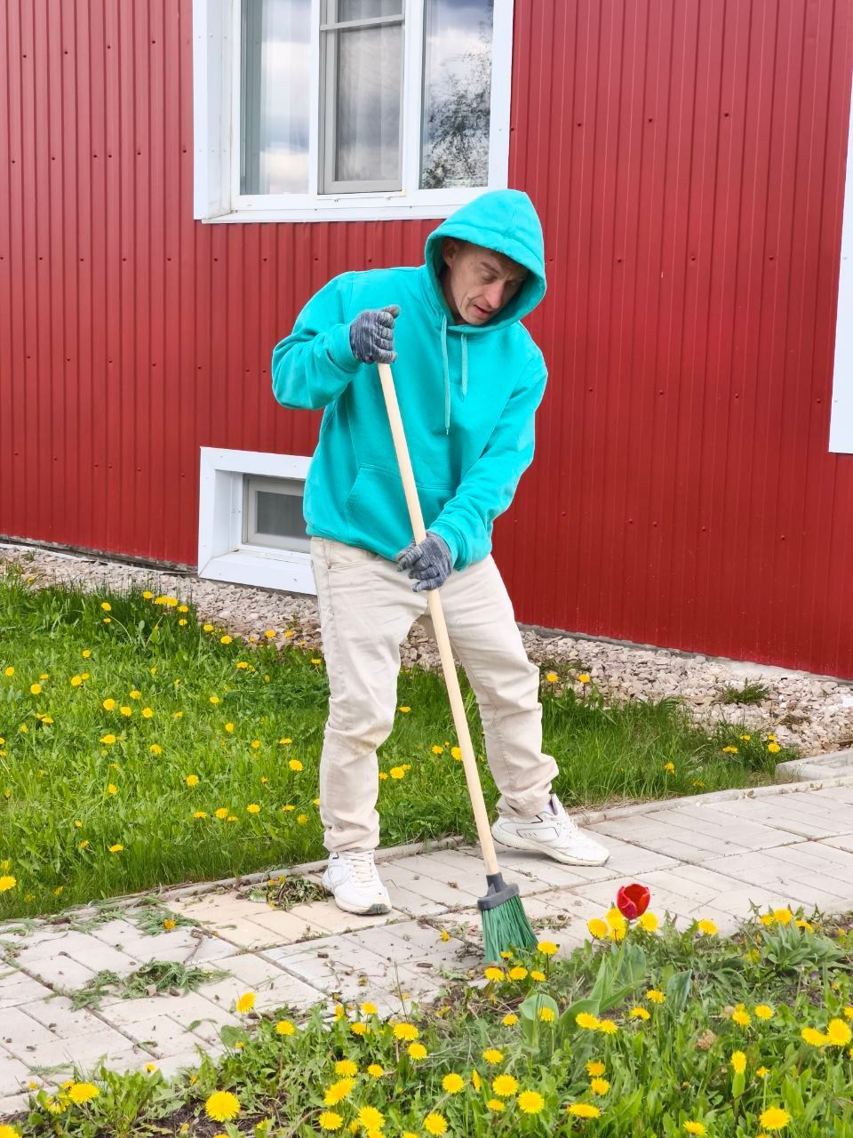 Person in turquoise hoodie using a broom to clean a sidewalk beside a red building and flowers.