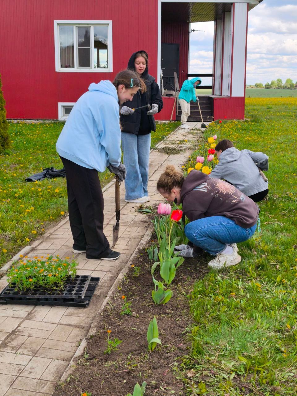 People planting flowers in a garden beside a red house under overcast skies