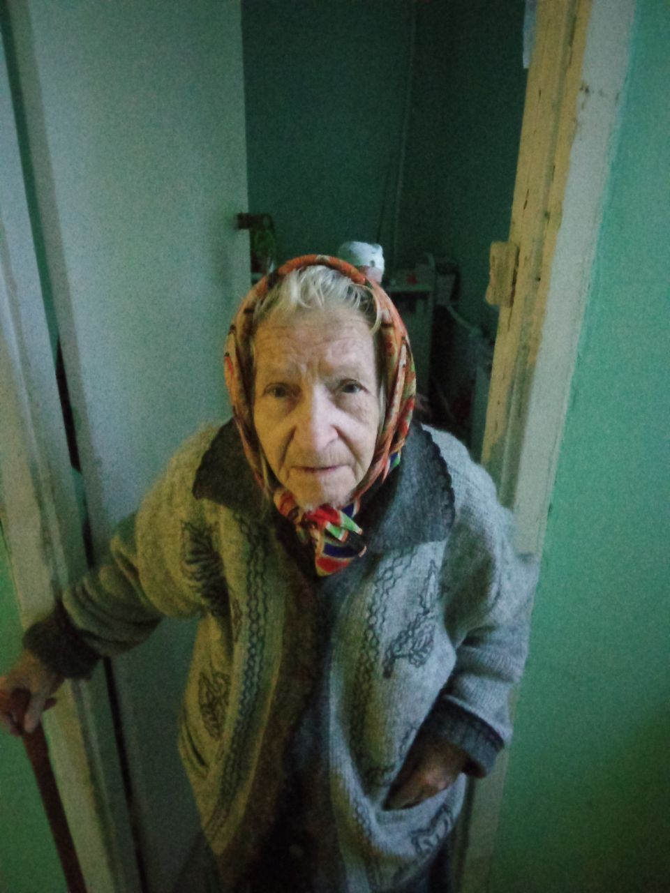 Elderly person in a gray sweater and red headscarf standing in a doorway, holding the doorframe.