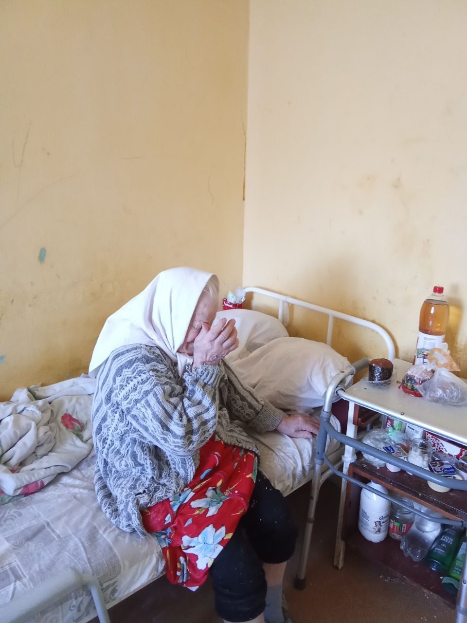 Person sitting on a bed in a small room, covering their face with a hand beside a bedside table.