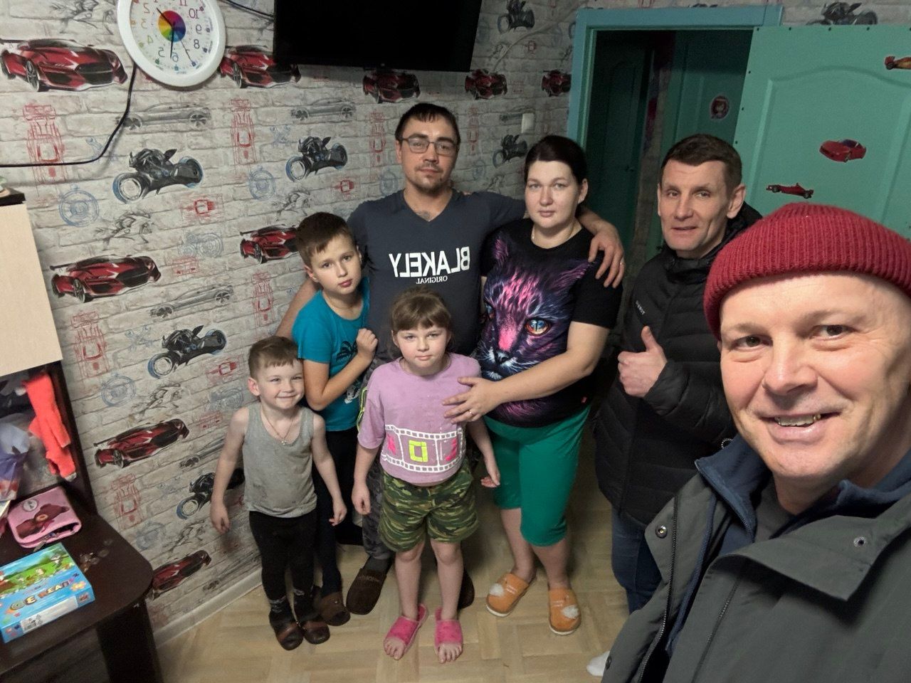 Family posing for a selfie in a room with car-pattern wallpaper and a TV on the wall.