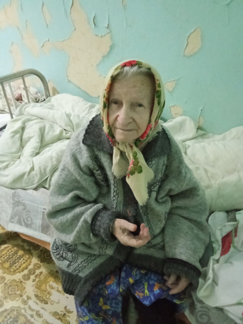 An elderly woman in a headscarf and coat sits on a bed in a worn room, holding an object in her hands.
