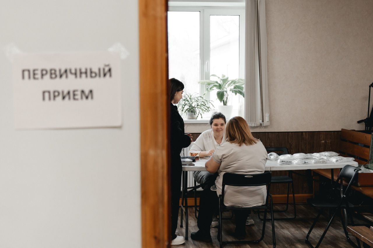 Reception room with people seated at a table, and a sign reading “Initial appointment” in Russian.