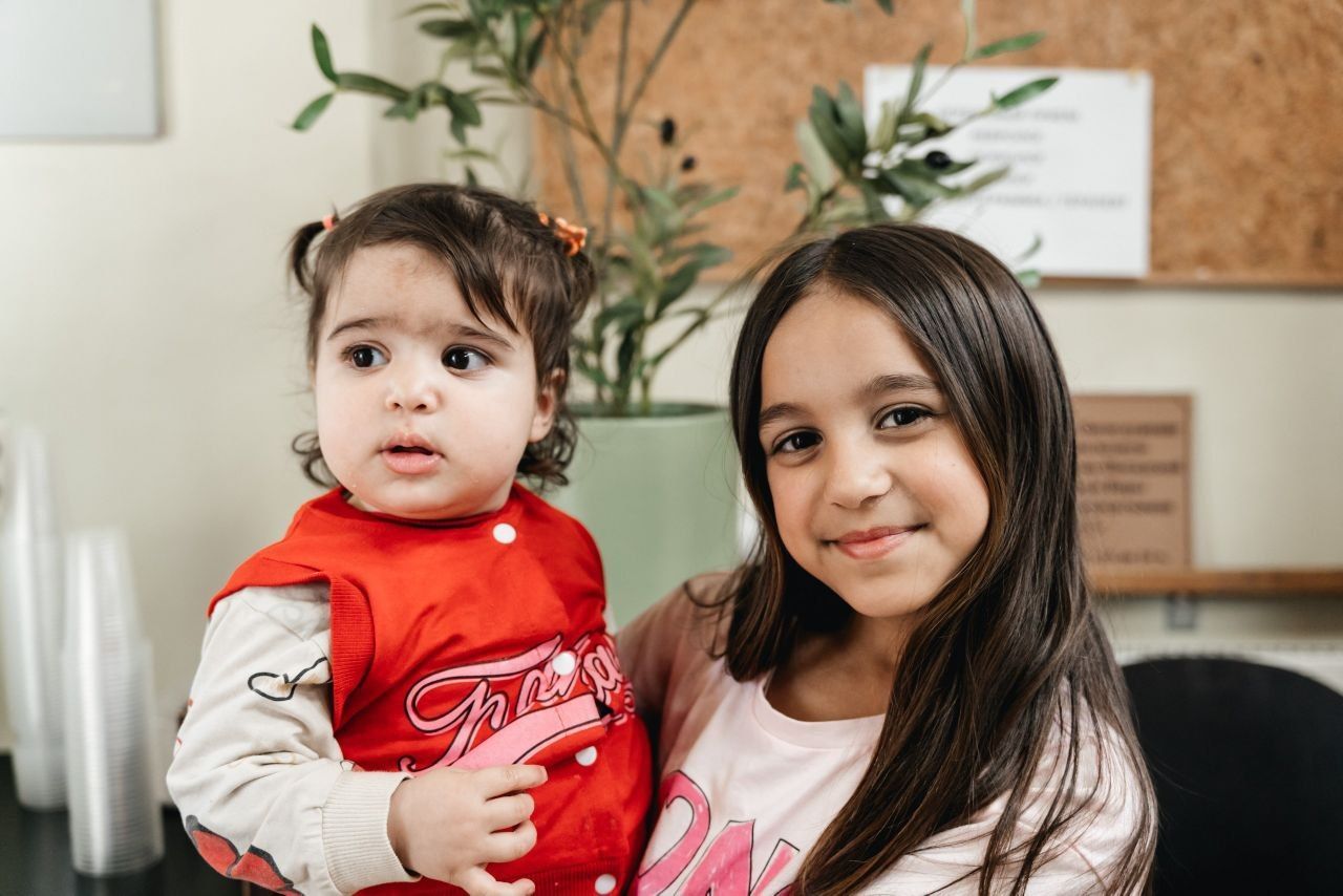 Two children posing indoors, one holding the other, with a soft smile and a potted plant in the background.