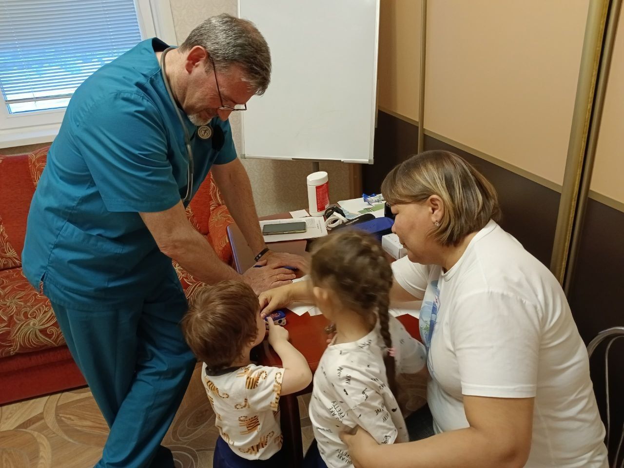 Two healthcare workers examine a young child in a clinic room, with another child nearby.