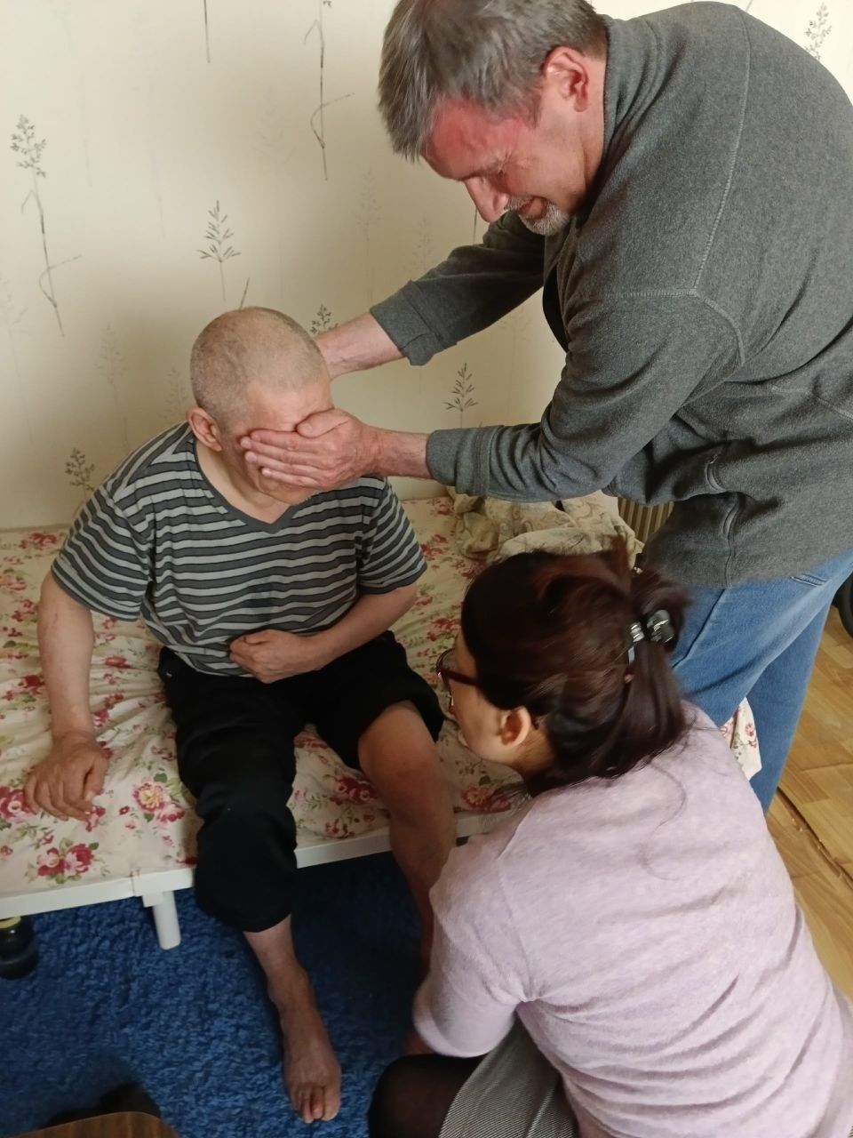 Three people care for a seated person in a striped shirt on a couch, indoors.