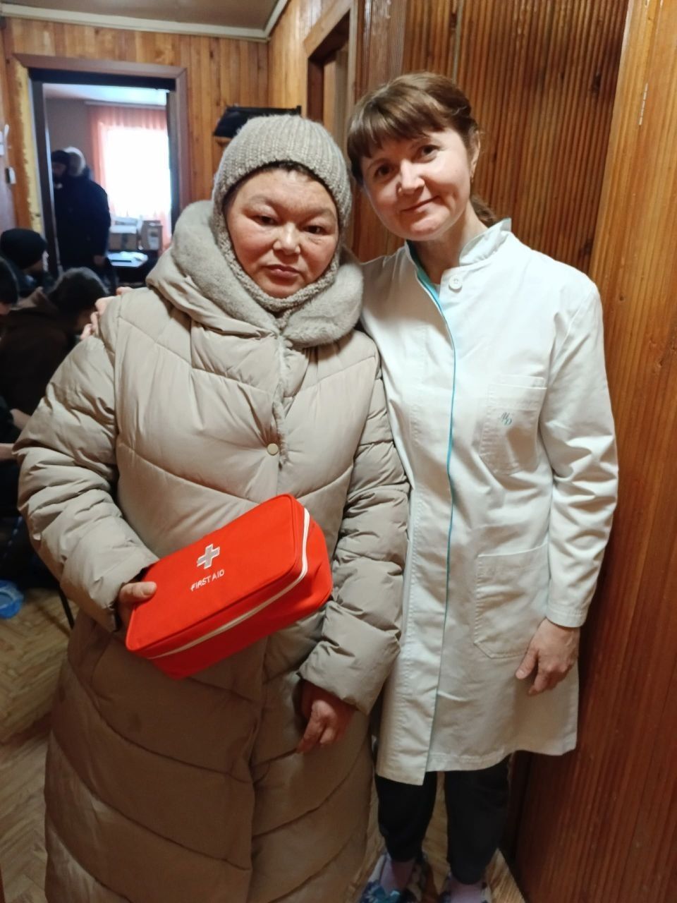 Two women stand indoors, one holding a red box and both smiling at the camera.